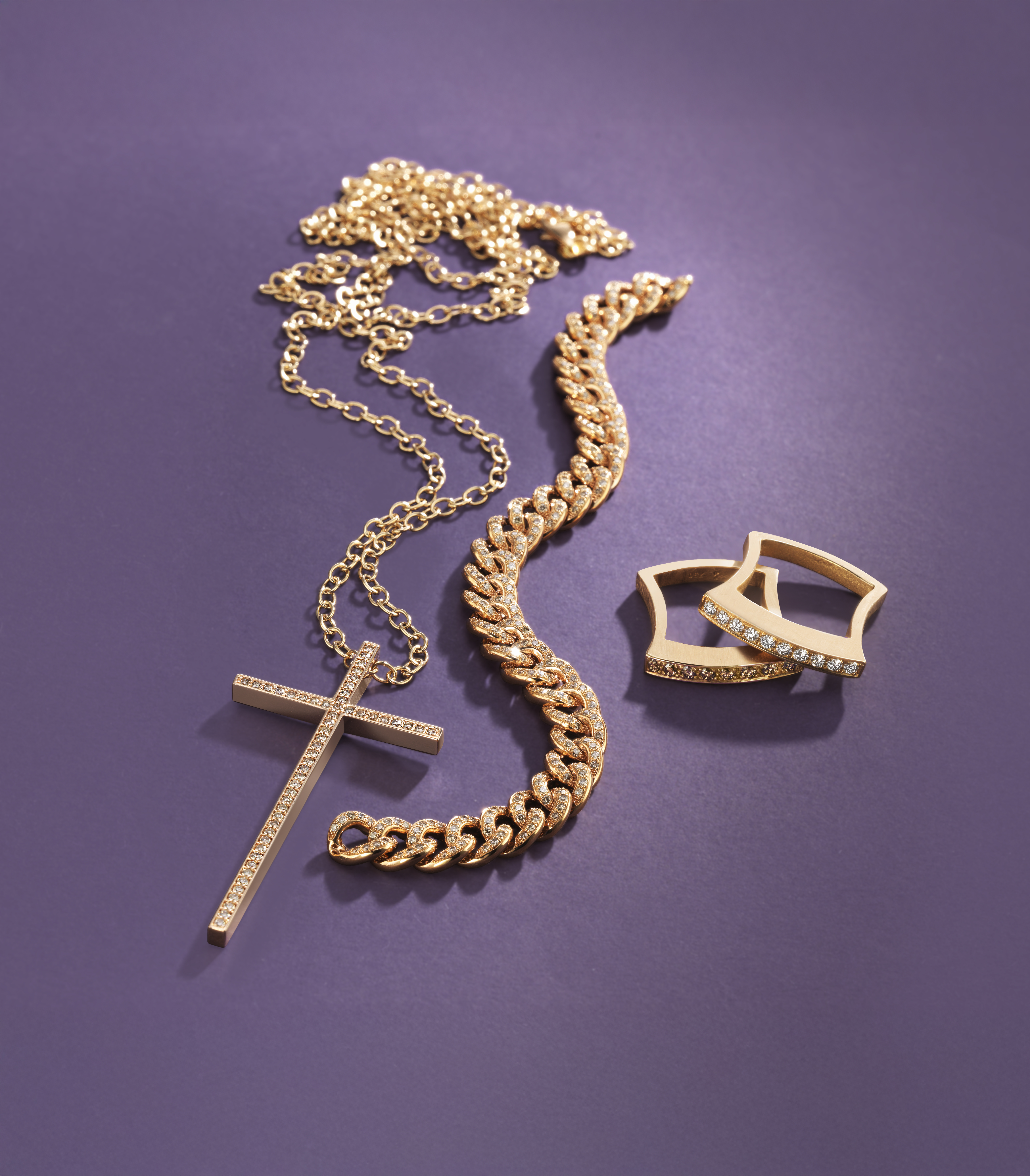 Eine feine goldene Kette mit Kreuzanhänger, ein goldenes Armband und zwei Ringe auf violettem Hintergrund